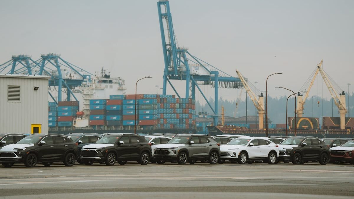 Eine Reihe geparkter Autos in einem geschäftigen Industriehafen mit Kranen und Containern
