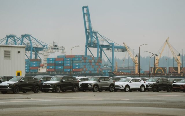 Eine Reihe geparkter Autos in einem geschäftigen Industriehafen mit Kranen und Containern