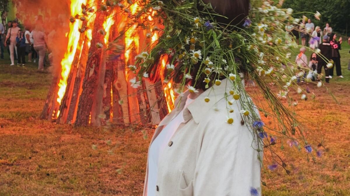 Eine lebendige Festivalszene mit einem großen Lagerfeuer und einer Person mit Blumenkranz im Freien