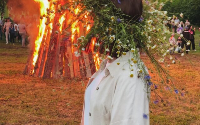 Eine lebendige Festivalszene mit einem großen Lagerfeuer und einer Person mit Blumenkranz im Freien