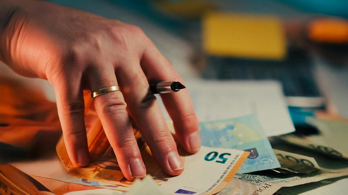 Eine Hand hält Euro-Banknoten über einem Schreibtisch mit Finanzdokumenten und einem Stift