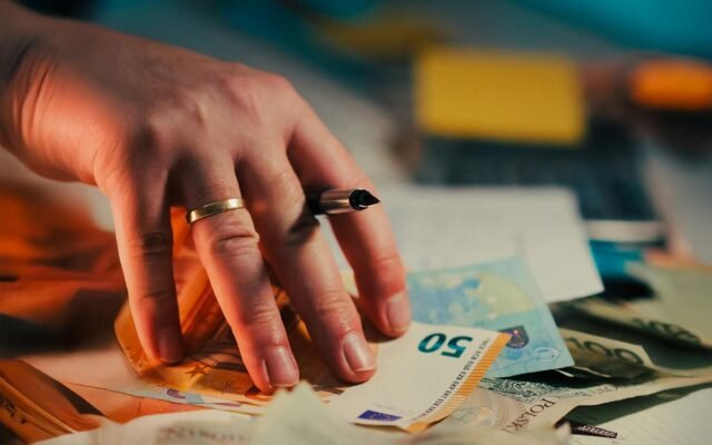 Eine Hand hält Euro-Banknoten über einem Schreibtisch mit Finanzdokumenten und einem Stift