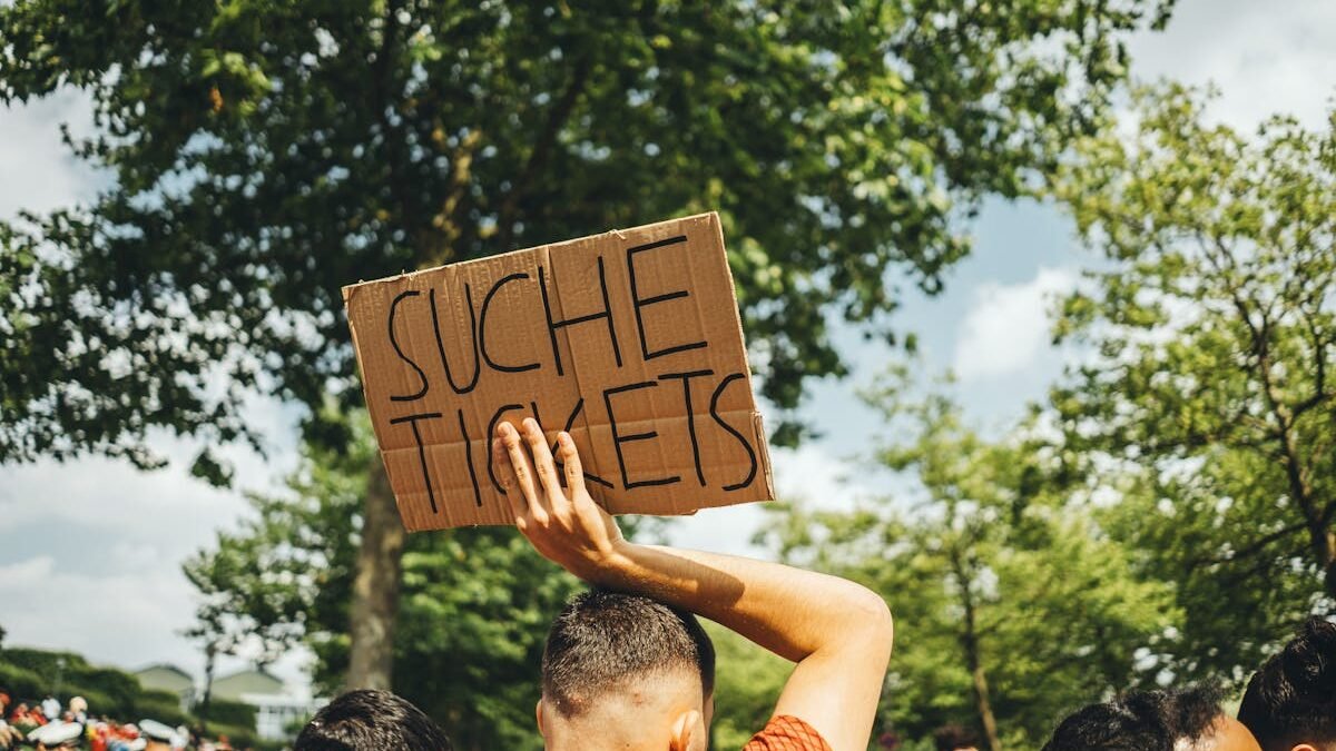 Ein Mann in einer Menschenmenge hält bei einer Veranstaltung im Freien ein Schild mit der Aufschrift „Suche Tickets“