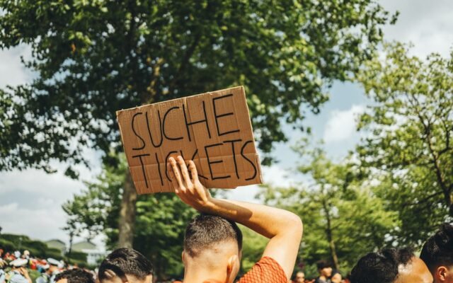 Ein Mann in einer Menschenmenge hält bei einer Veranstaltung im Freien ein Schild mit der Aufschrift „Suche Tickets“